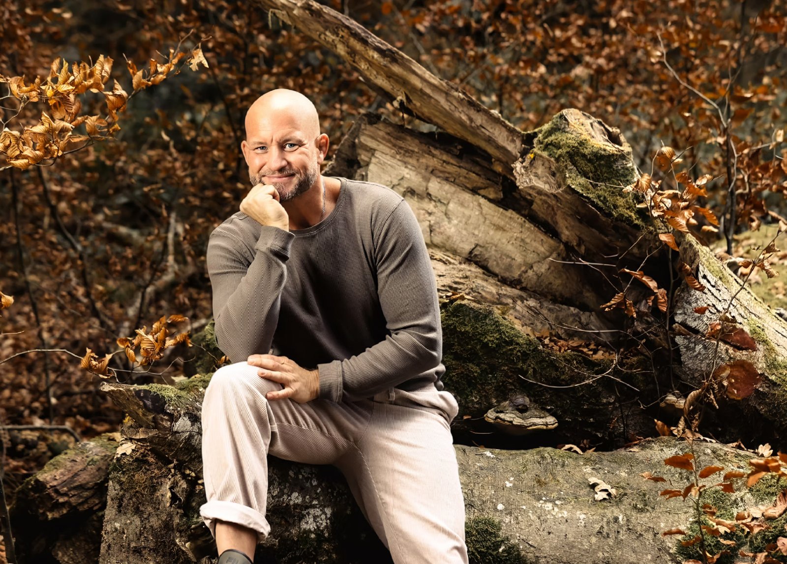 André Walberer, Hypnose- und Energiecoach in Schiefling am Wörthersee, sitzt lächelnd mit der Hand am Kinn auf einem moosigen Baumstamm im Herbstwald