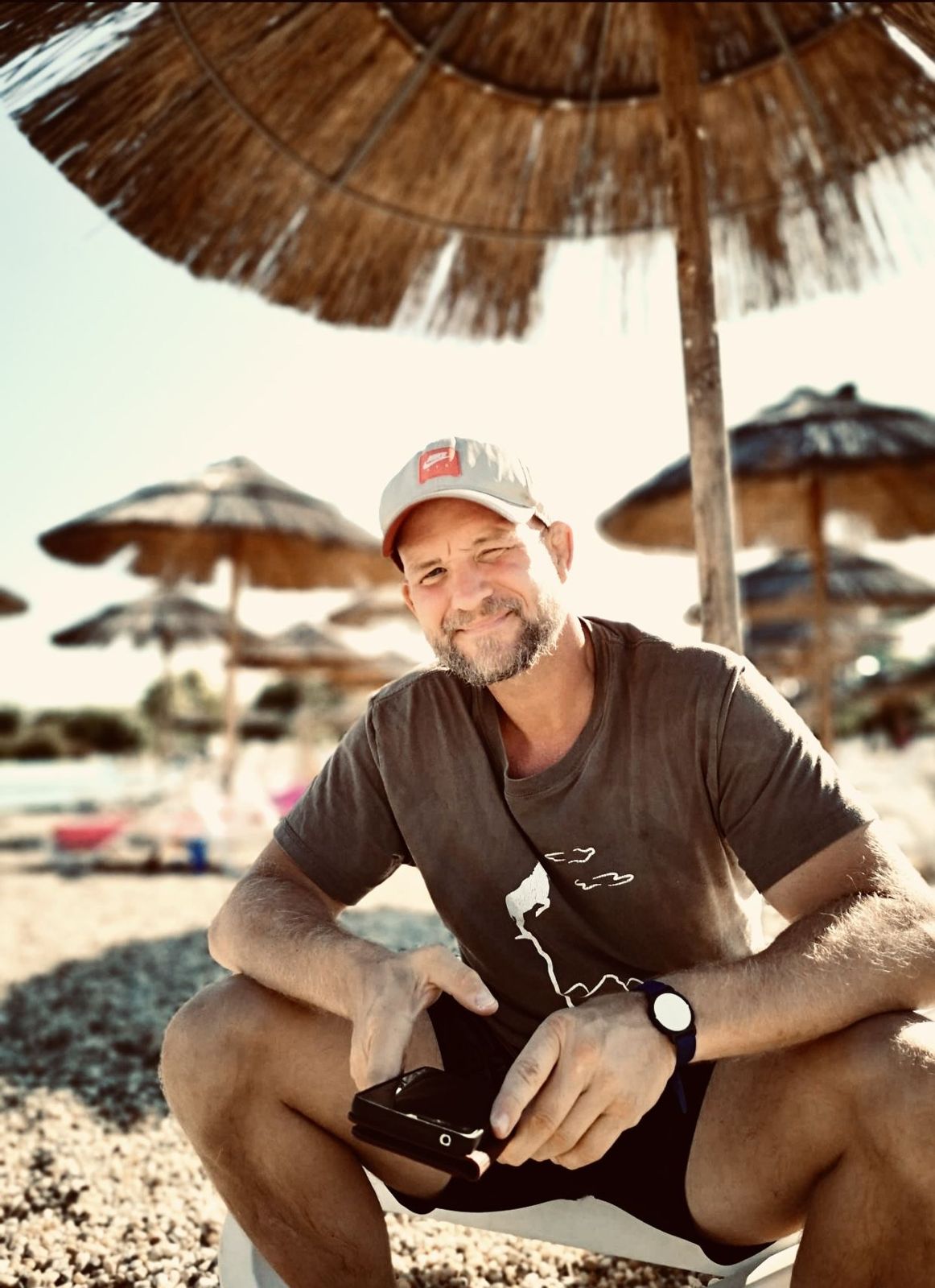André Walberer, Hypnose- und Energiecoach, sitzt lächelnd mit Kappe am Strand unter Sonnenschirmen