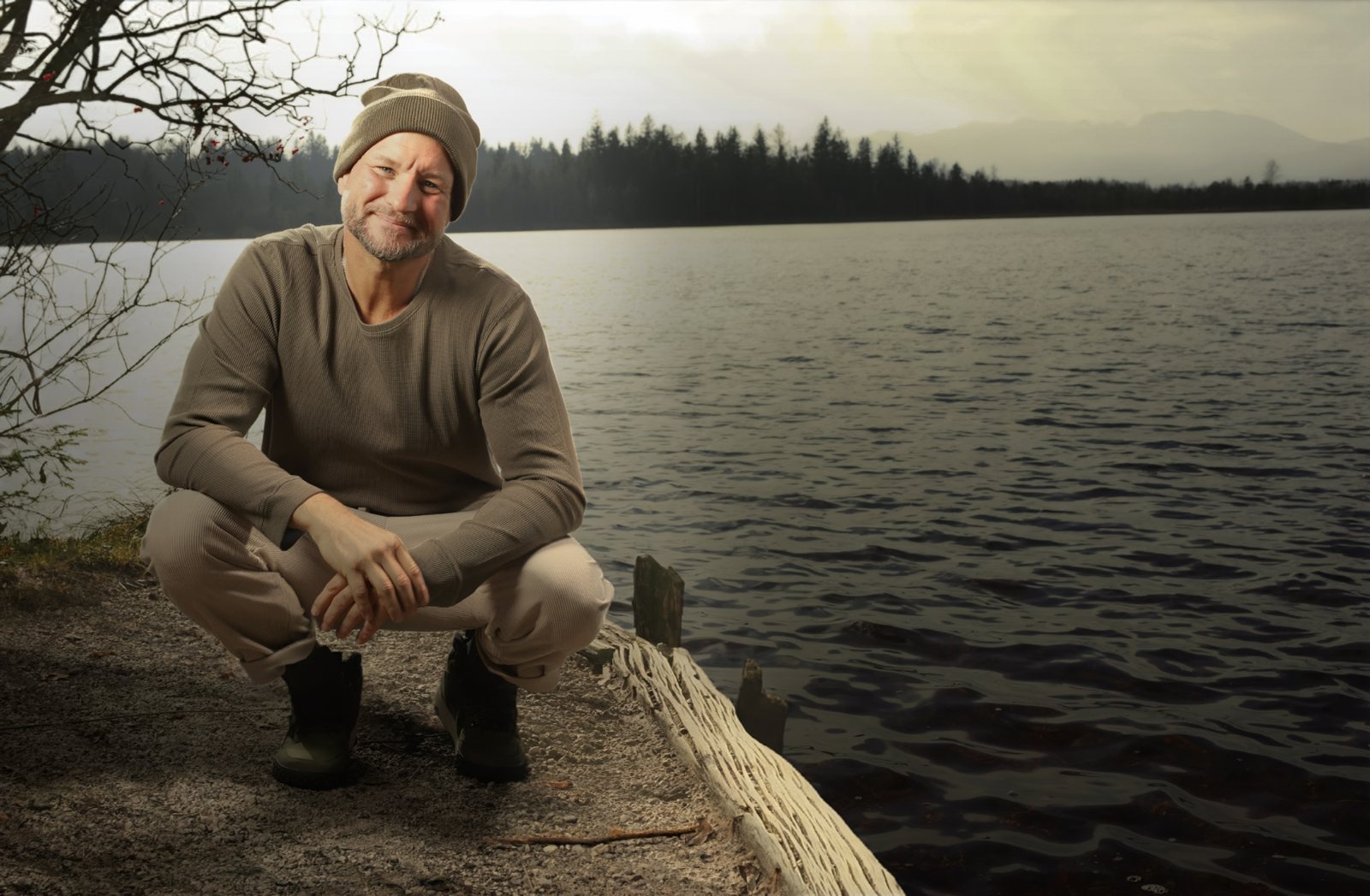 Bewusstseins- und Naturcoaching am See in Kärnten: André Walberer hockt lächelnd am Seeufer, dahinter Wald und Berge